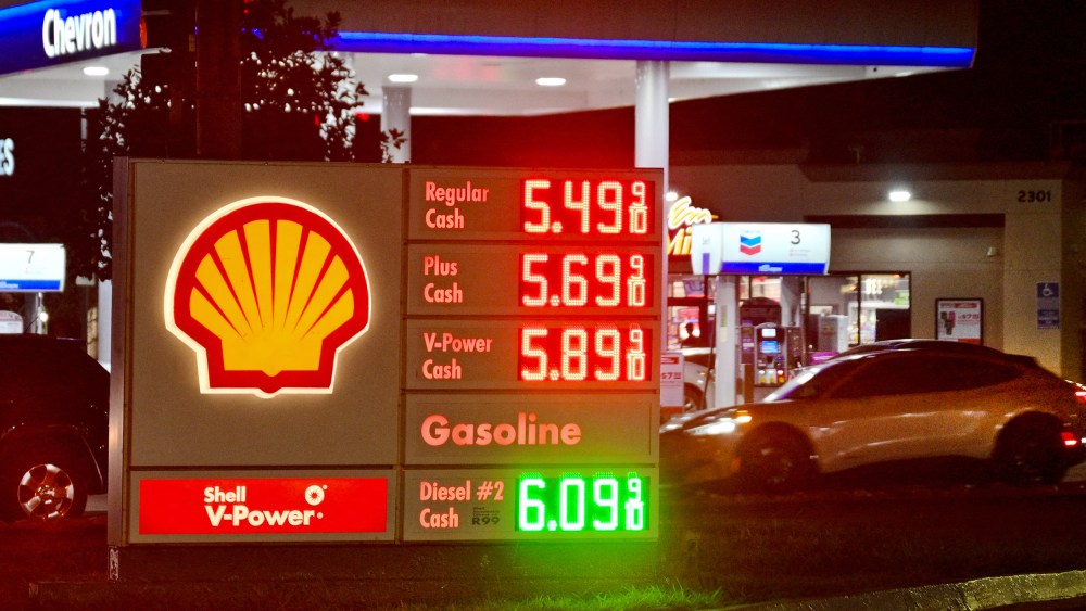 A gas station sign on the ground shows fuel prices while a car pulls into to get gas.