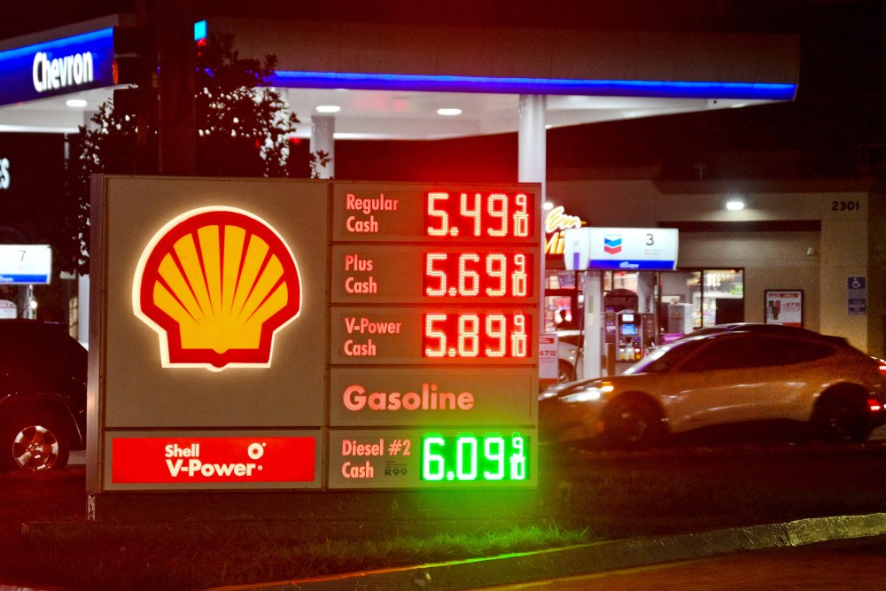 A gas station sign on the ground shows fuel prices while a car pulls into to get gas.